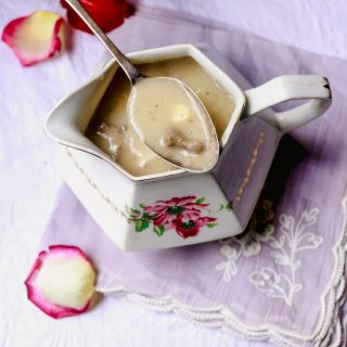 Giblet Gravy in a rose stenciled serving dish with spoon of gravy laying on top and rose petals surrounding it on lavender tablecloth