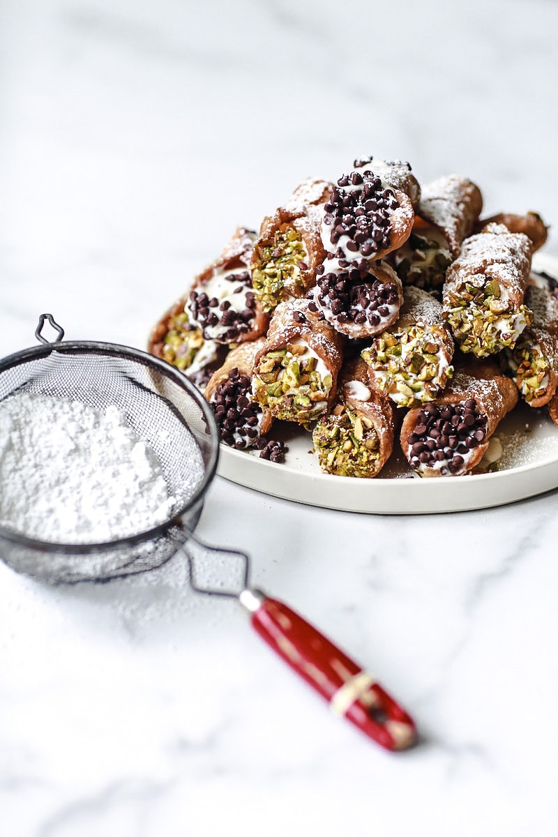 Cannoli on a round plate stacked up with chocolate and pistachios on white marble with powdered sugar sprinkle