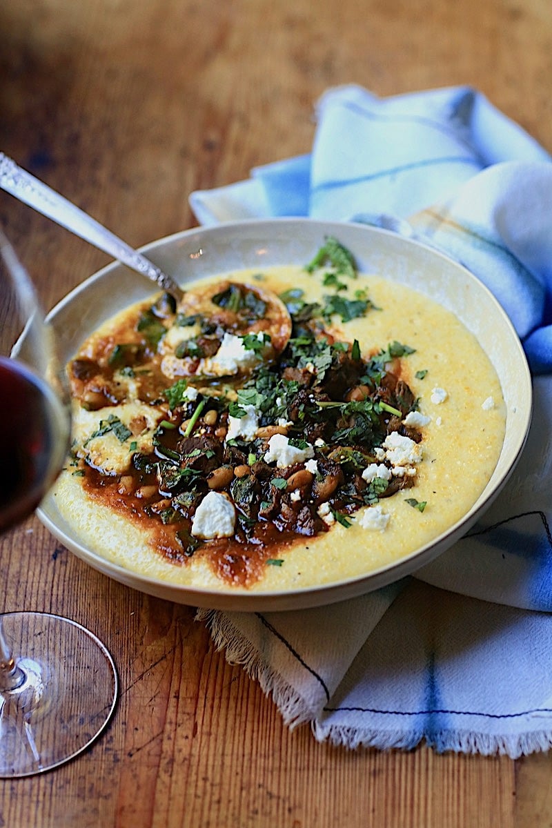 Bow of Cheesy Grits with meat, chickpeas, gravy, herbs, and goat cheese with a spoon and glass of wine on wooden surface