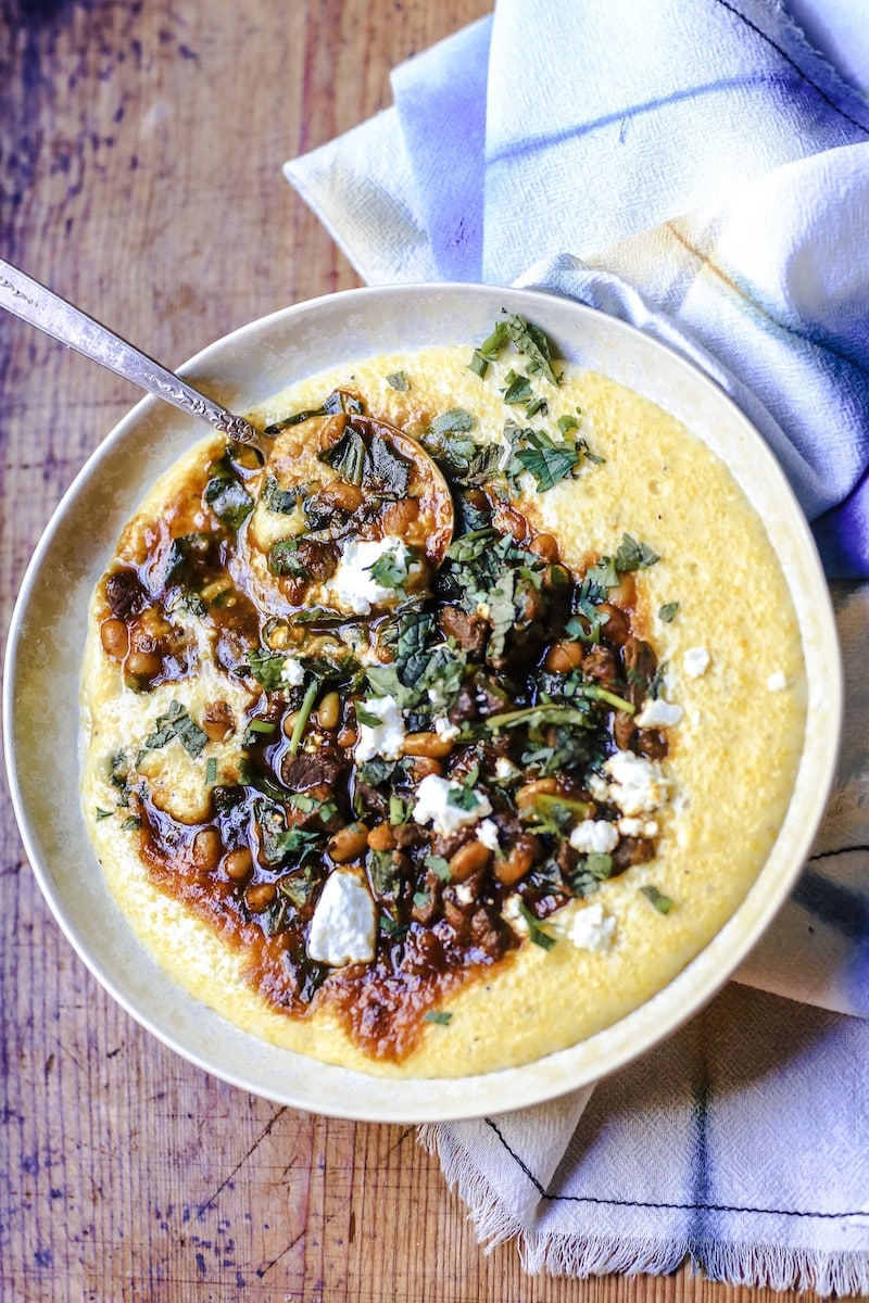 Bowl of Persian Stew with venison, beans, collards, spices and herbs, and topped with goat cheese in a bowl of cheesy grits with folded napkin on wooden table