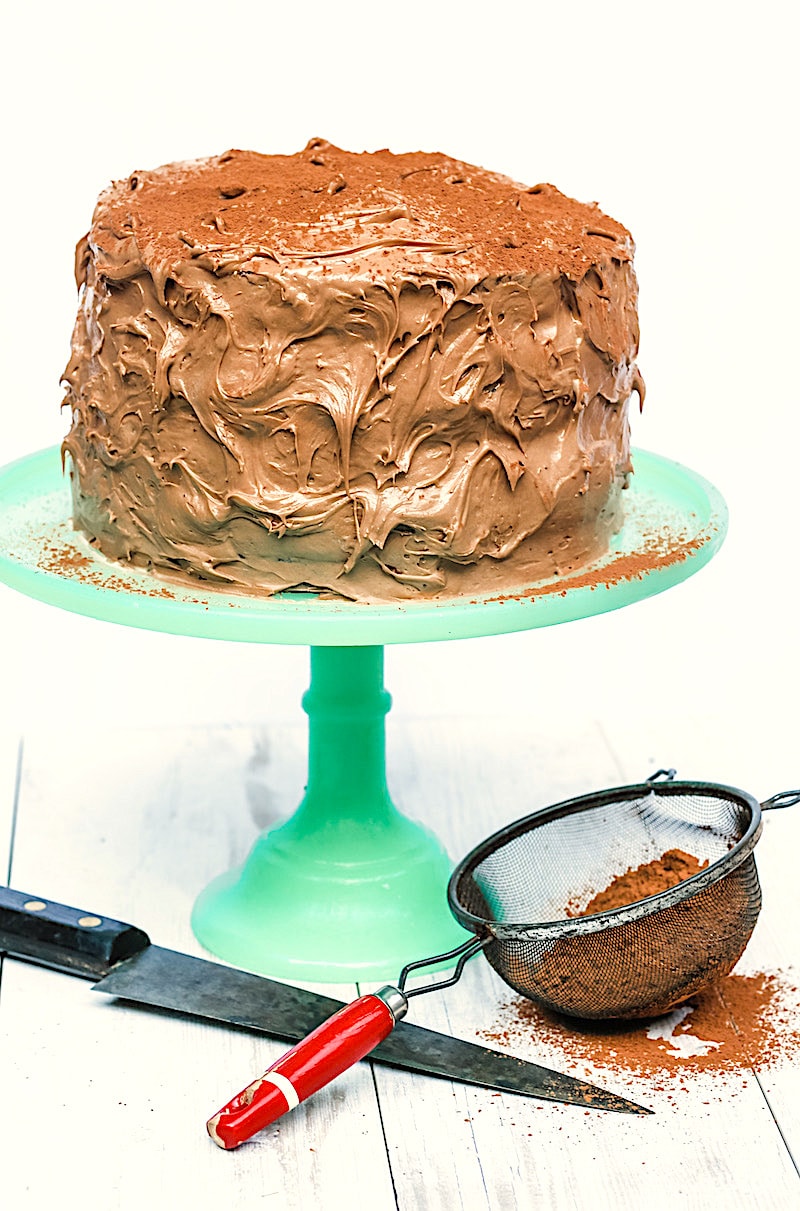 4 layer cappuccino cake on green cake stand with knife in front of it along with cocoa powder in a sieve 
