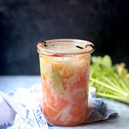 Jar of Pickled Daikon and Carrots with a blue and white napkin and radish leaves behind the jar