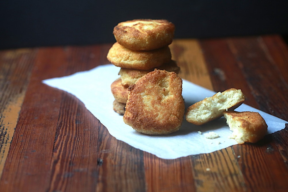 Horizontal photo of stacked fried cornbread shaped like discs with one propped up on the stacked cornbread and one broken open to see the inside over parchment on wood and black background.
