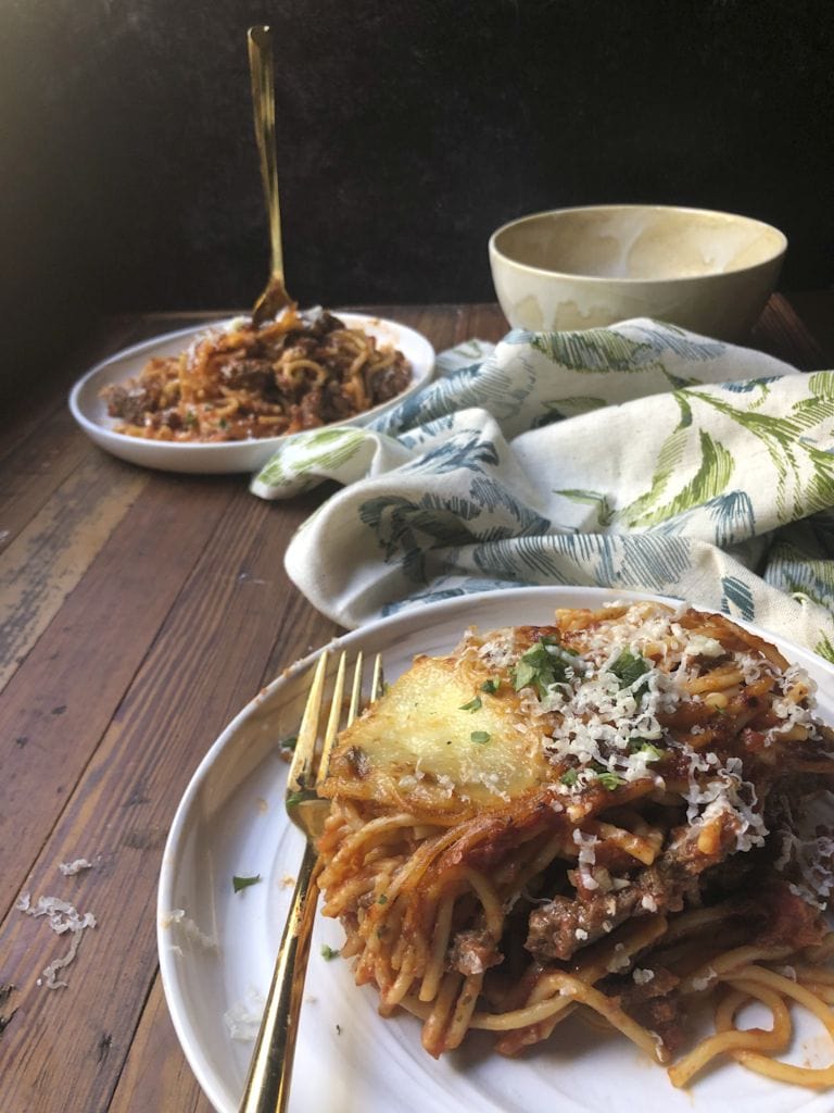 closeup of serving of baked spaghetti with fork on white plate with another plate behind it and linen floral napkin