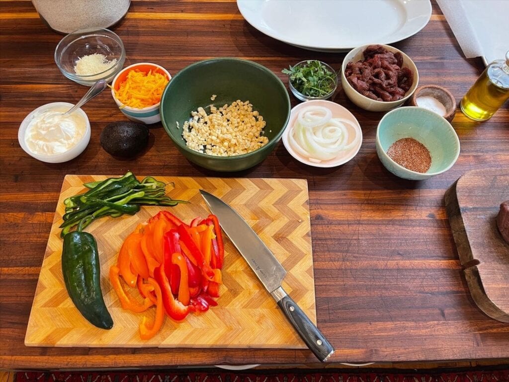 sliced red and green peppers and poblano pepper on wood cutting board, bowls of corn and other ingredients in background