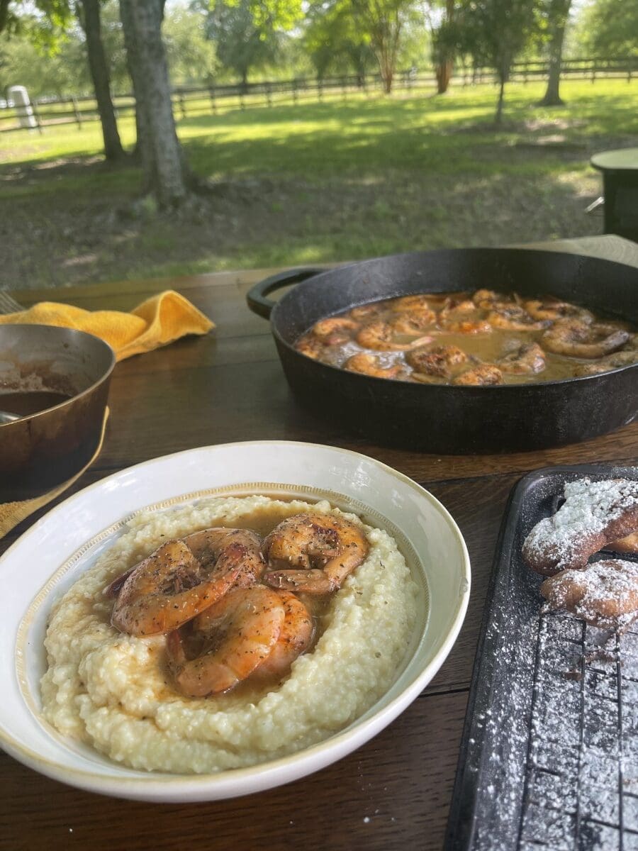 feast of BBQ shrimp over cheesy grits next to powdered beignets with cast-iron skillet of shrimp in background