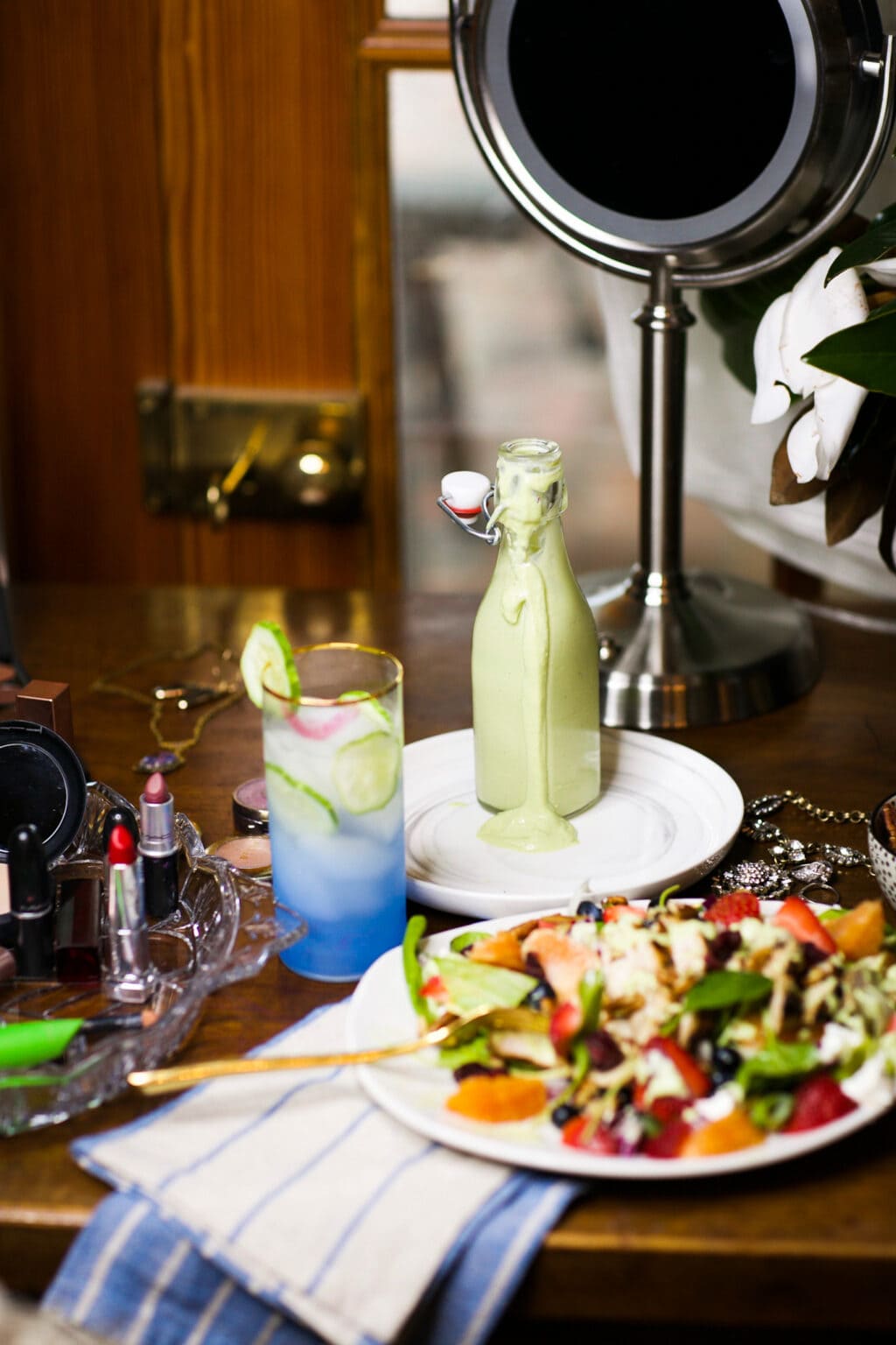 avocado dressing, green goddess style, in glass bottle behind white plate full of power salad with chicken and a blue beverage with ice. Salad is next to a tray with lipsticks.