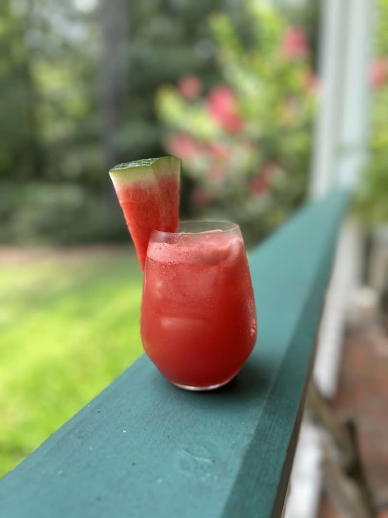 glass of red watermelon lemonade drink with watermelon wedge, resting on green porch railing