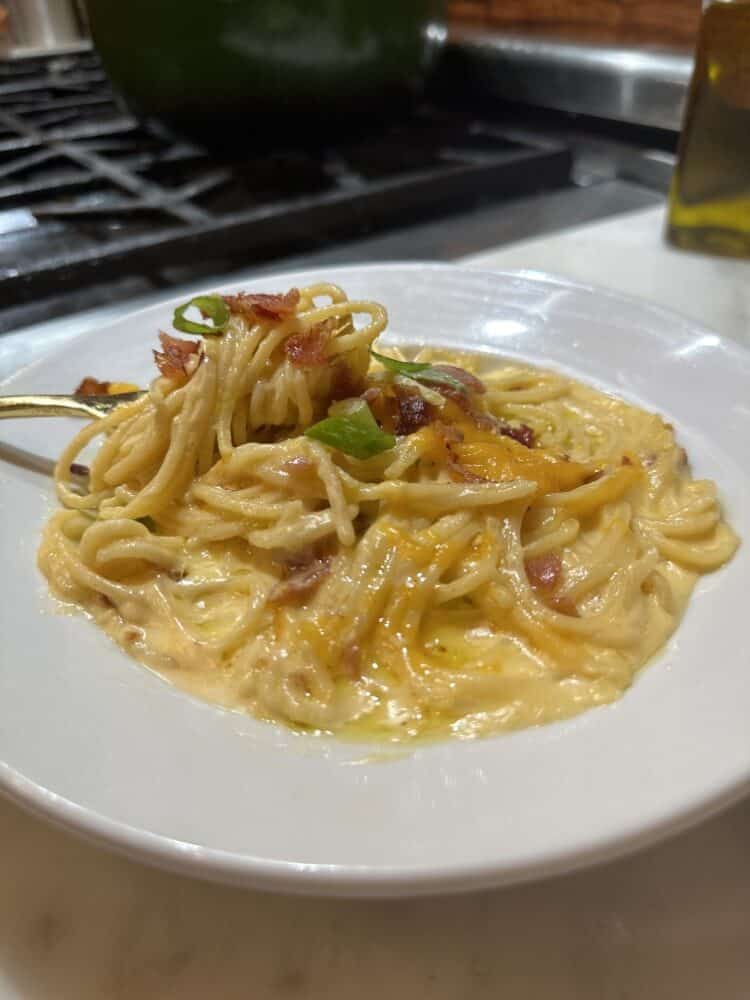white shallow bowl with gruyere mac and cheese spaghetti topped with bacon and green onions - fork with twirled pasta is resting on side of plate
