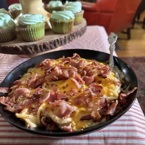 Large plate of New Orleans mac and cheese spaghetti, topped with bacon pieces, on red-striped tablecloth with green-frosted cupcakes in background