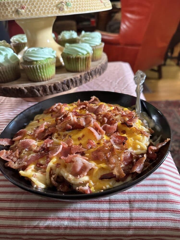 Large serving plate of New Orleans mac and cheese spaghetti, topped with bacon pieces, on red-striped tablecloth with green-frosted cupcakes in background
