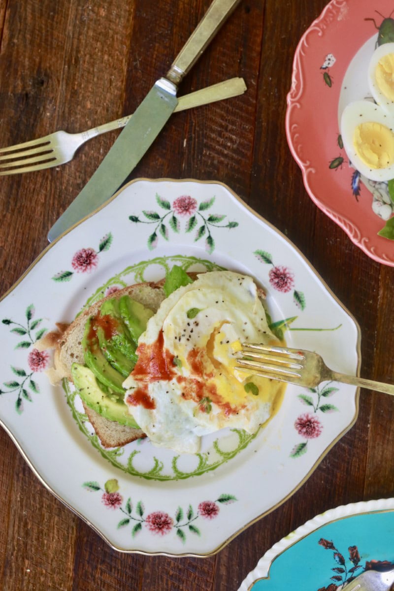 fried eggs with avocado toast on white plate with floral design, fork resting on yolk of eggs